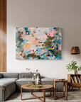 An abstract painting with a colorful paint splatter pattern in shades of blue, pink, beige, and green, displayed in a modern living room setting above a gray sofa.