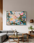 An abstract framed painting with splashes of pink, blue, green, and cream hangs on a light gray wall above a gray sectional sofa in a modern living room setting.