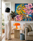 A woman in a white outfit stands in a bright living room holding a potted plant, facing a large colorful floral painting on the wall. The room includes a beige sofa, a small wooden side table with an orange lamp, and a blue bin.