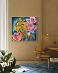 A colorful abstract floral painting hanging on a warm beige wall in a modern room setting with a brown woven chair, blue rug, and a plant in the foreground.