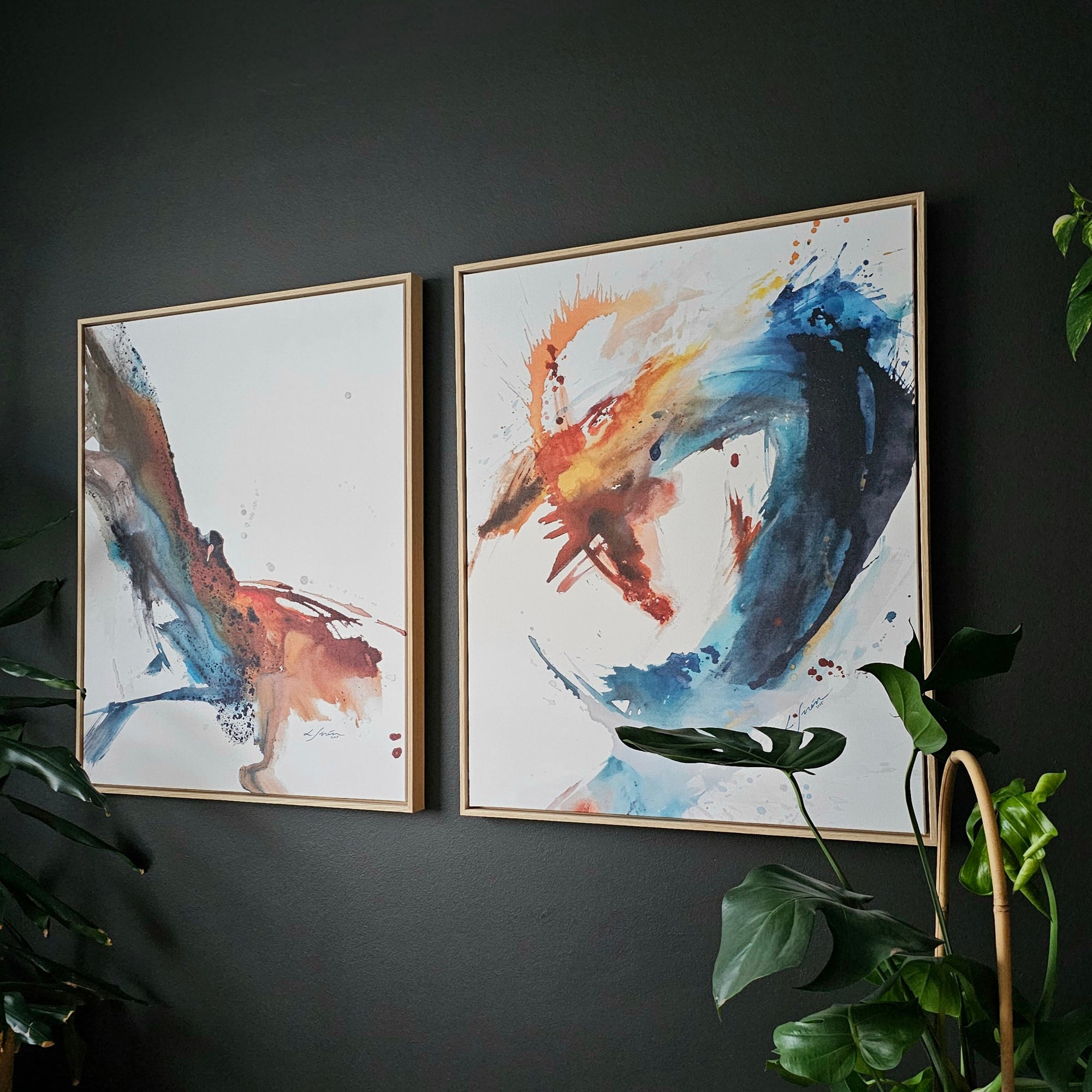 Two abstract art prints by Lotta Sirén on a dark wall with green plants around.