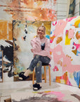Lotta Sirén sitting on a stool in front of a colorful abstract painting wearing a light pink sweatshirt with abstract graphic on the front