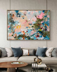 An abstract canvas painting with vibrant blue, pink, green, and gold brushstrokes, displayed on a textured light gray wall above a modern beige sofa in a styled living room.