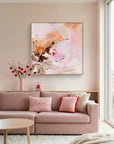 An abstract painting with pink, peach, white, and brown brushstrokes hanging on a beige wall above a pink sofa in a bright, modern living room.