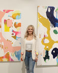 Lotta Sirén standing between two abstract paintings with vibrant colors on a wall. Strawberry Kisses abstract art piece on the left of her.