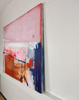 A side profile of an abstract painting with pink, blue, and brown colors on a white wall.