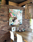 An abstract canvas painting with pink, blue, and white brushstrokes hanging on a rustic red brick wall above a wooden dining table with white chairs in a modern loft setting.