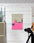 Camera on a tripod and an abstract painting "No Matter" with pink and beige colors against a white wooden studio wall.