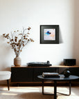 A framed abstract art print with blue, white, and peach tones hangs on a white wall in a modern living room setting above a black media console with a turntable, speaker, and dried flowers.