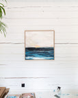Framed artwork of a seascape on a white wooden wall with art supplies on a table below by Finnish abstract artist Lotta Sirén