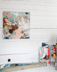 An abstract canvas painting with a multicolor paint-splattered design hangs on a white wood-paneled wall in an artist's studio. A rolled canvas and other art pieces are visible on the floor, along with a metal paint storage cart.