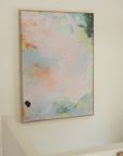An abstract framed canvas art print with soft pastel brushstrokes in pink, blue, green, and peach, hanging on a warm beige wall.