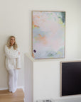 Sara Lindholm in a white knit sweater and white pants stands beside a large framed abstract canvas print on a white wall in a bright, modern room with light wood flooring.