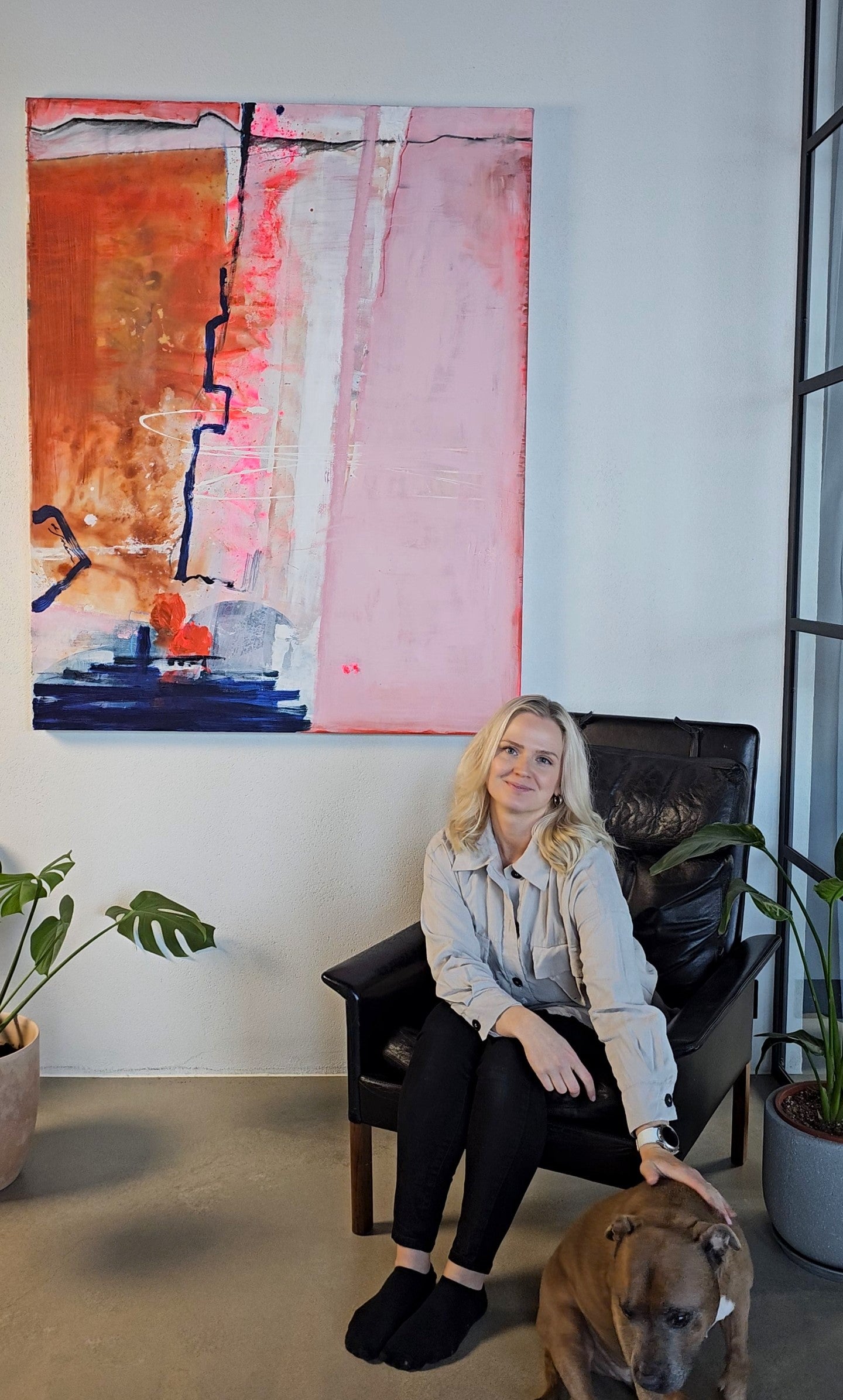 Lotta Sirén sitting on a chair with a dog in a room with large abstract painting on thee wall and plants.