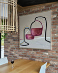 An abstract painting of wine glasses a with pink, purple, black, and blue brushstrokes on a cream background, hanging on a brick wall above a wooden dining table with a glass of red wine.
