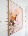 An abstract framed painting with pink, peach, brown, and white brushstrokes, hanging on a white wall.
