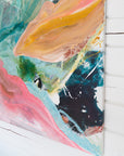 An abstract canvas painting with expressive brushstrokes in teal, pink, peach, orange, black, and white, shown hanging on a white wood-paneled wall.