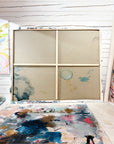 A large four-panel wooden canvas frame set against a white wood-paneled wall. A paint-splattered palette lies on the floor in the foreground.