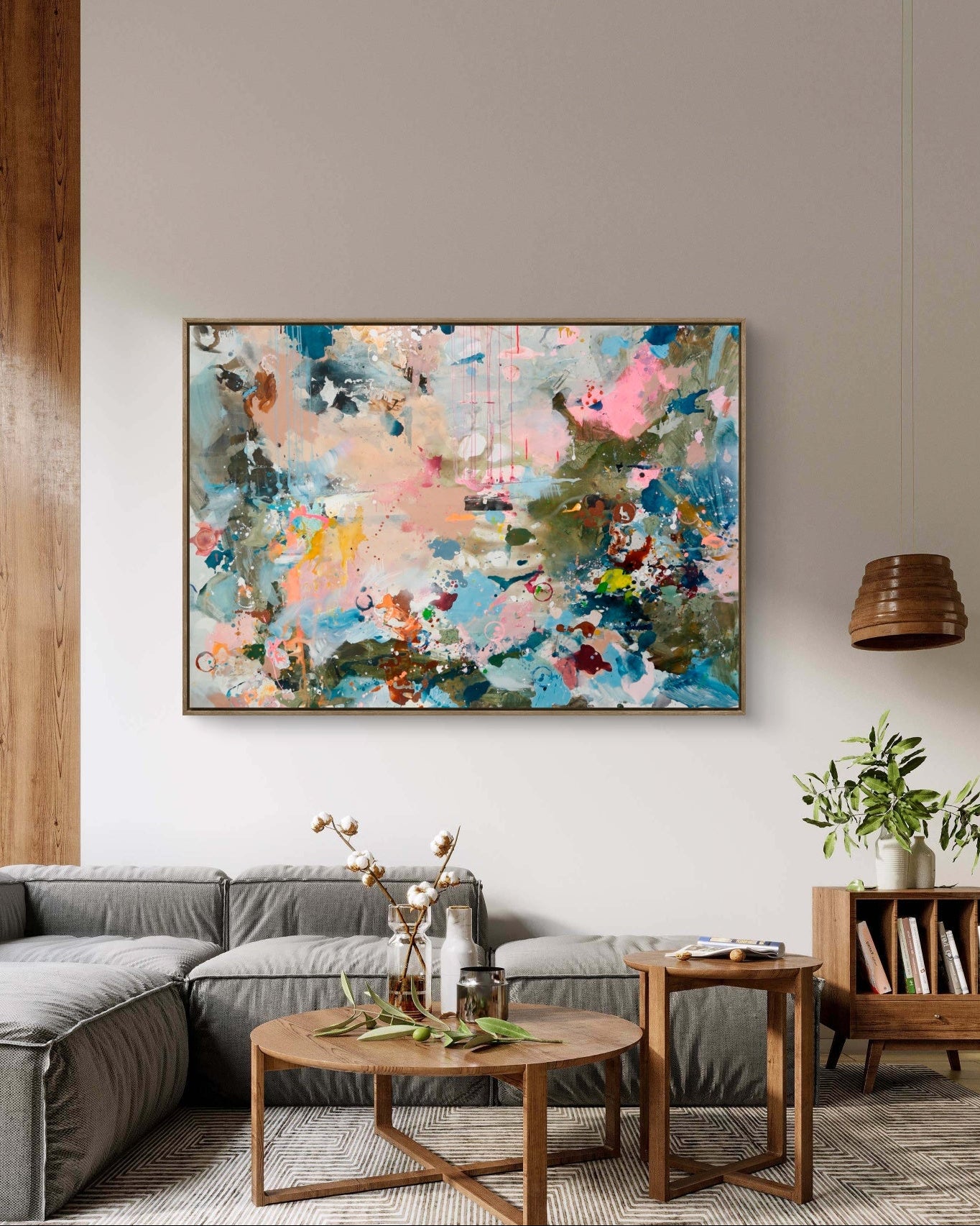 An abstract framed painting with splashes of pink, blue, green, and cream hangs on a light gray wall above a gray sectional sofa in a modern living room setting.
