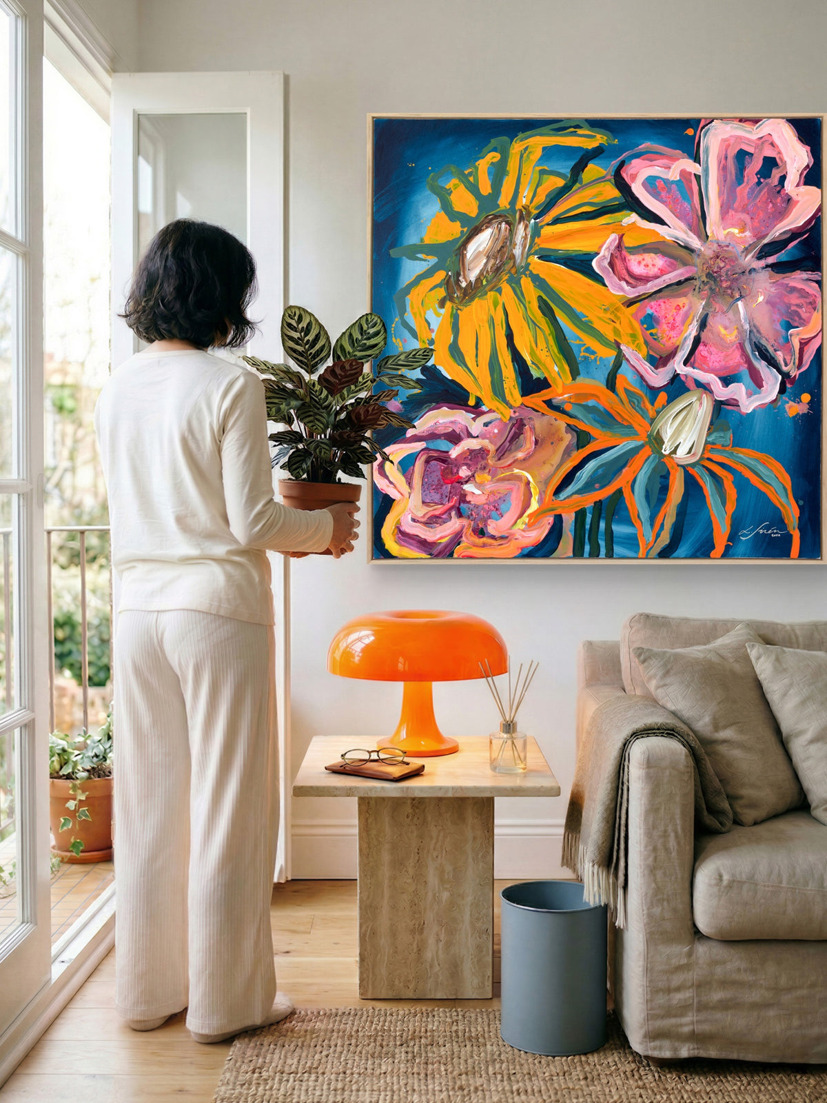 A woman in a white outfit stands in a bright living room holding a potted plant, facing a large colorful floral painting on the wall. The room includes a beige sofa, a small wooden side table with an orange lamp, and a blue bin.