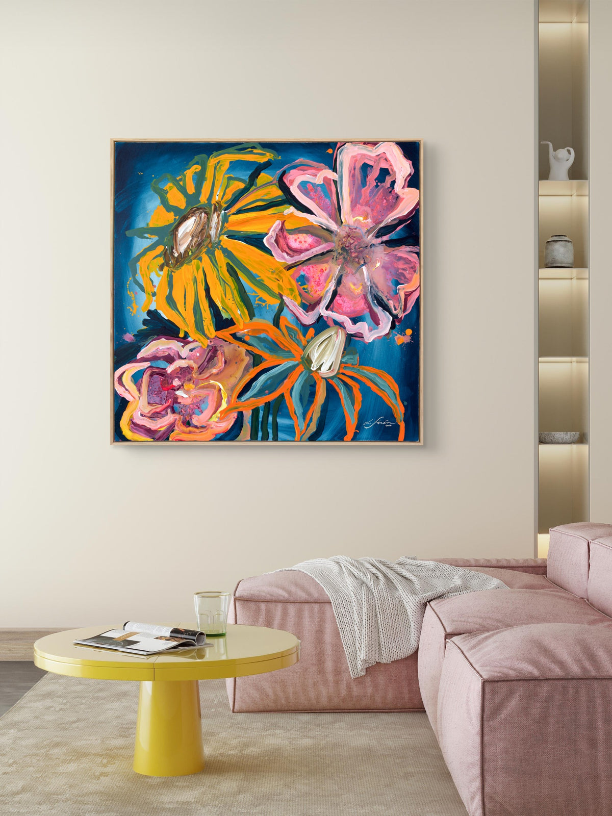 A colorful abstract floral painting with pink, yellow, and orange flowers on a dark blue background, displayed on a beige wall above a pink sofa in a modern living room setting.