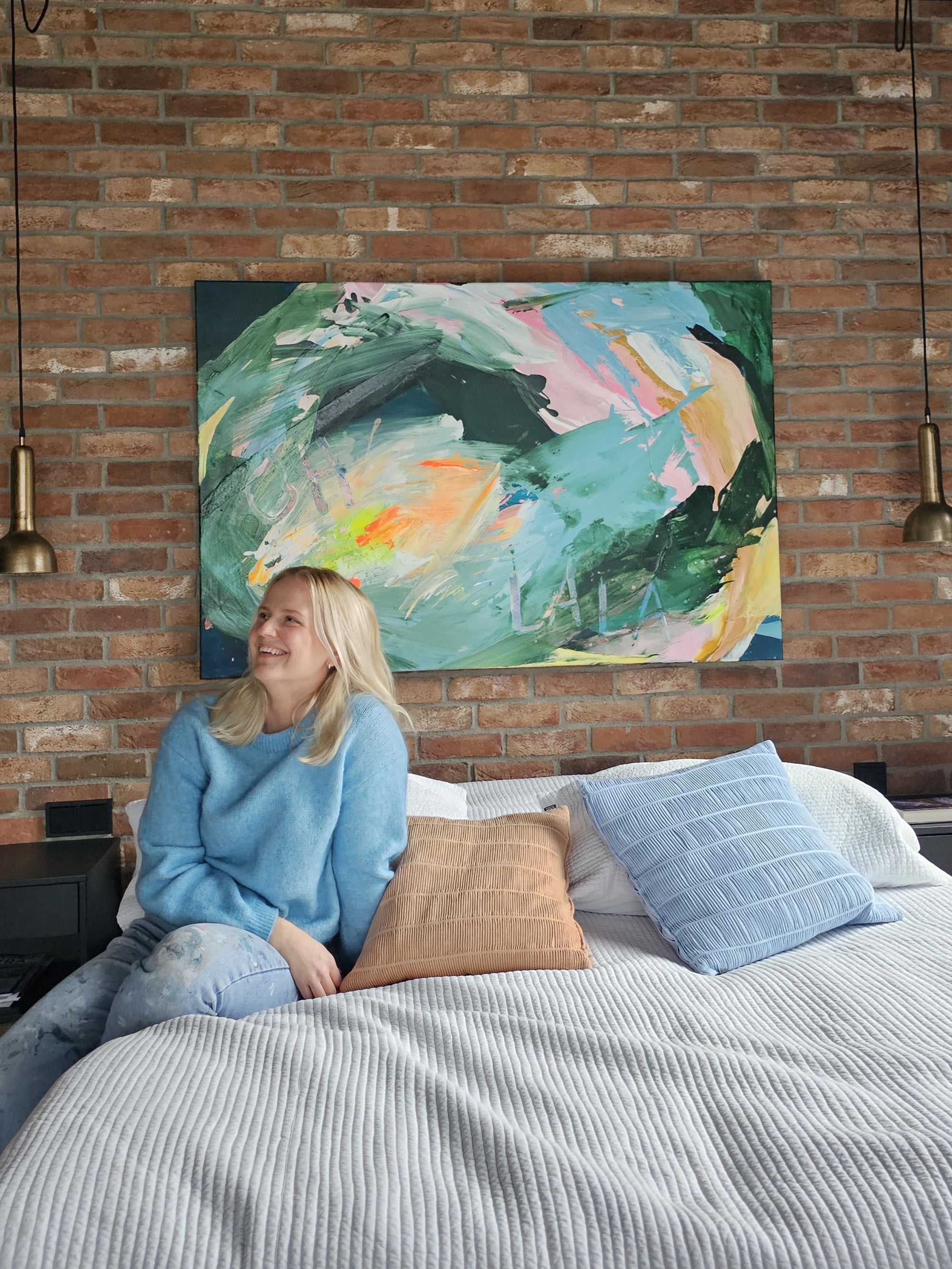 Lotta Sirén sitting on a bed in a room with a colorful green abstract painting on the wall.