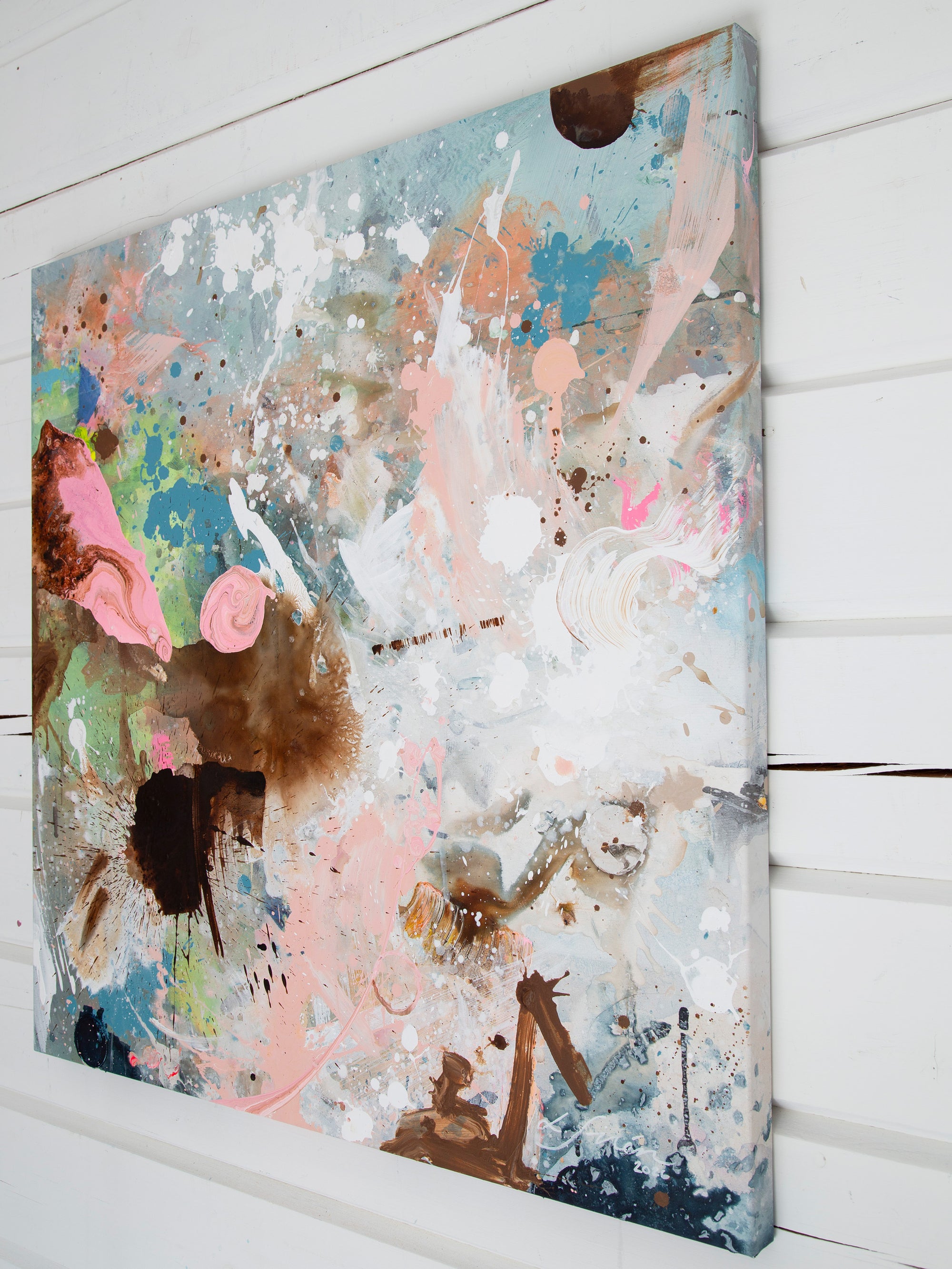An abstract canvas painting with splatters, drips, and brushstrokes in pastel pink, light blue, white, brown, and green, shown hanging on a white shiplap wall.