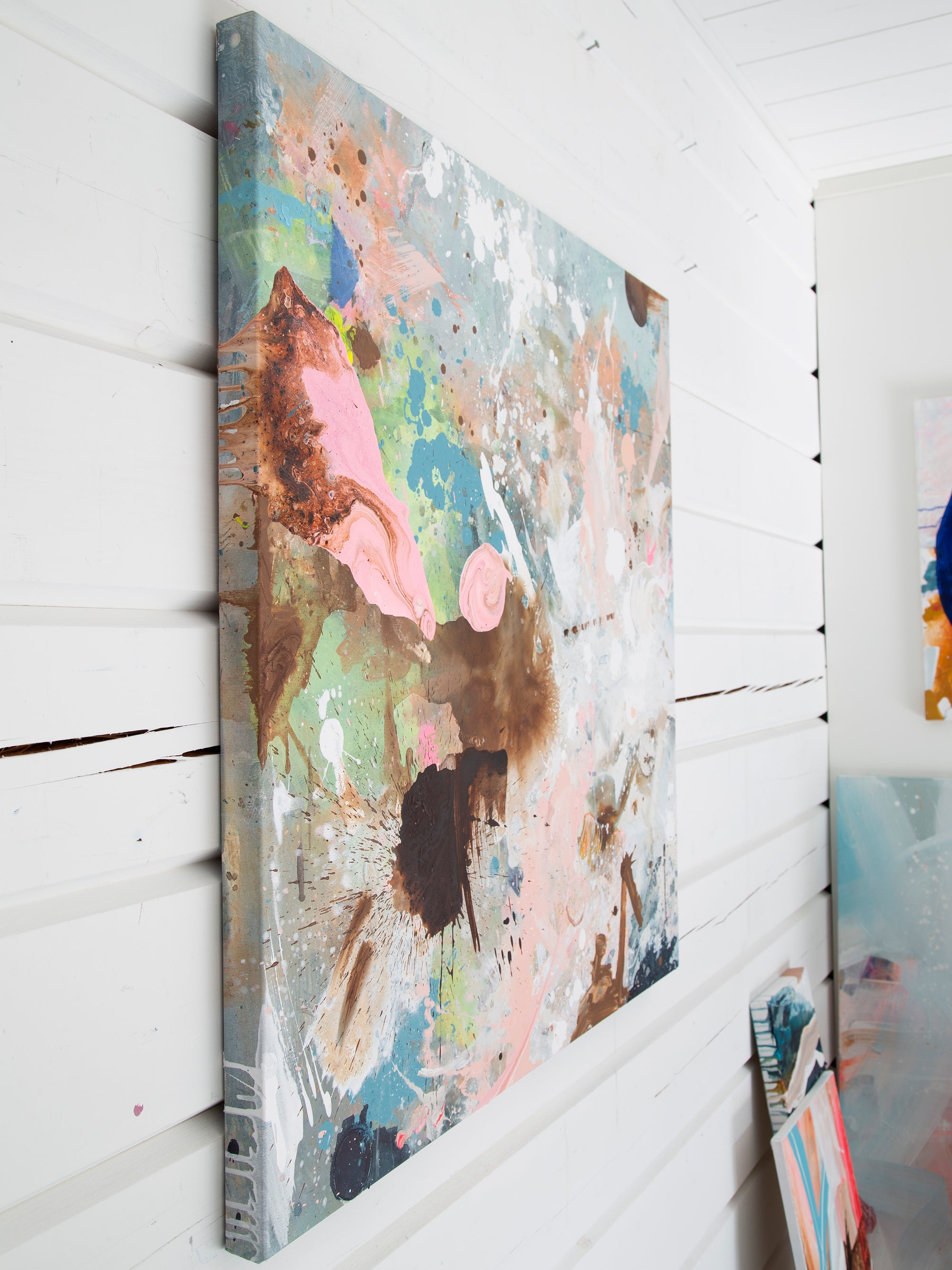 An abstract canvas wall art print with splatters and drips of pink, blue, brown, and white paint, shown hanging on a white paneled wall.