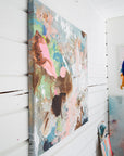 An abstract canvas wall art print with splatters and drips of pink, blue, brown, and white paint, shown hanging on a white paneled wall.