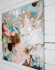 An abstract canvas painting with splatters, drips, and brushstrokes in pastel pink, light blue, white, brown, and green, shown hanging on a white shiplap wall.