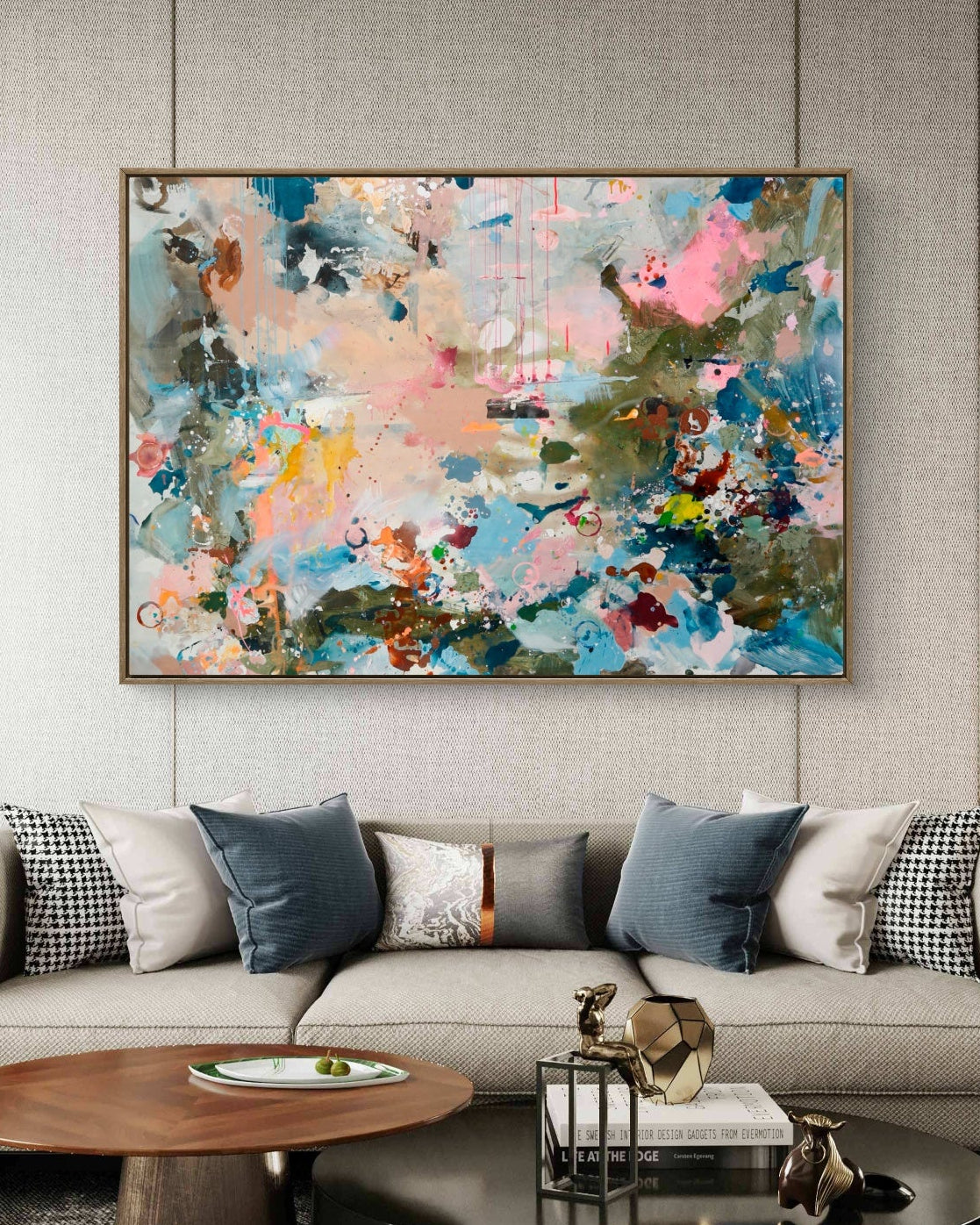 An abstract canvas painting with vibrant blue, pink, green, and gold brushstrokes, displayed on a textured light gray wall above a modern beige sofa in a styled living room.