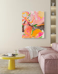 An abstract canvas painting featuring vibrant pink, orange, yellow, and green brushstrokes, displayed in a modern living room setting above a pink sofa and a yellow side table.