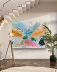 A large abstract canvas painting with vibrant splashes of turquoise, pink, orange, yellow, and green, displayed on a white wall in a modern home interior with a staircase, floor lamp, and potted plant.