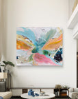 An abstract canvas wall art featuring a vibrant butterfly-like design in shades of pink, teal, yellow, orange, and green, displayed on a white wall in a modern living room setting.