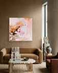 An abstract painting "I Need Space with" pink, peach, and brown brushstrokes hanging on a wooden wall in a modern living room setting with a tan sofa and stone coffee table.