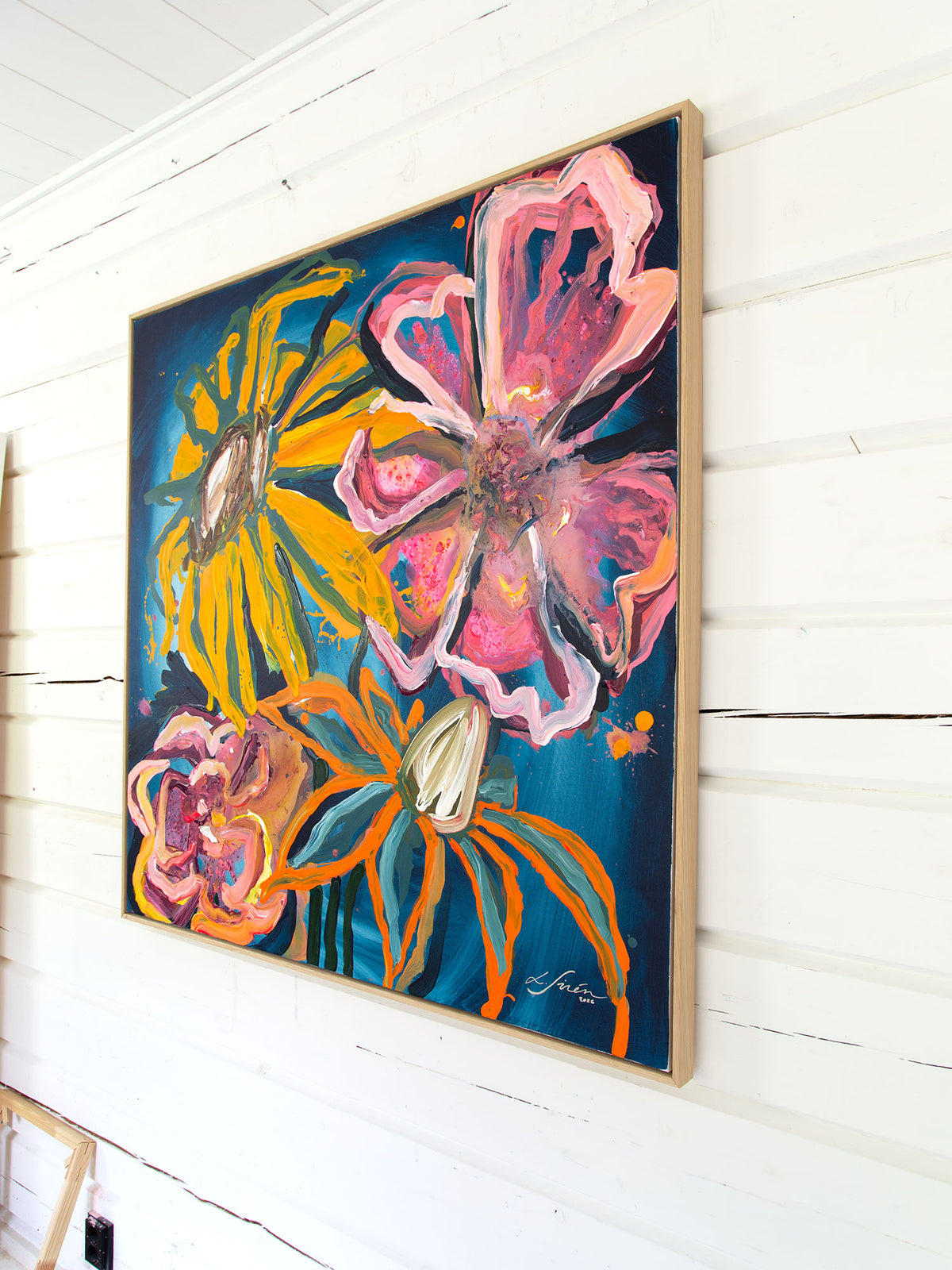 A framed abstract floral painting with bold pink, yellow, orange, and blue brushstrokes, hanging on a white wood wall. 