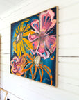 A framed abstract floral painting with bold pink, yellow, orange, and blue brushstrokes, hanging on a white wood wall. 