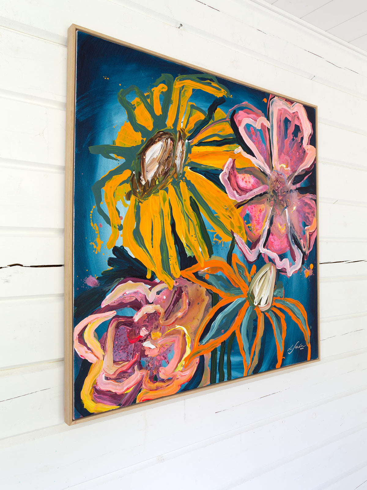 A framed abstract floral painting hanging on a white wood-paneled wall. The artwork features bold yellow, pink, orange, and blue flower-like forms on a dark blue background, enclosed in a light wood frame.