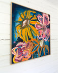 A framed abstract floral painting hanging on a white wood-paneled wall. The artwork features bold yellow, pink, orange, and blue flower-like forms on a dark blue background, enclosed in a light wood frame.