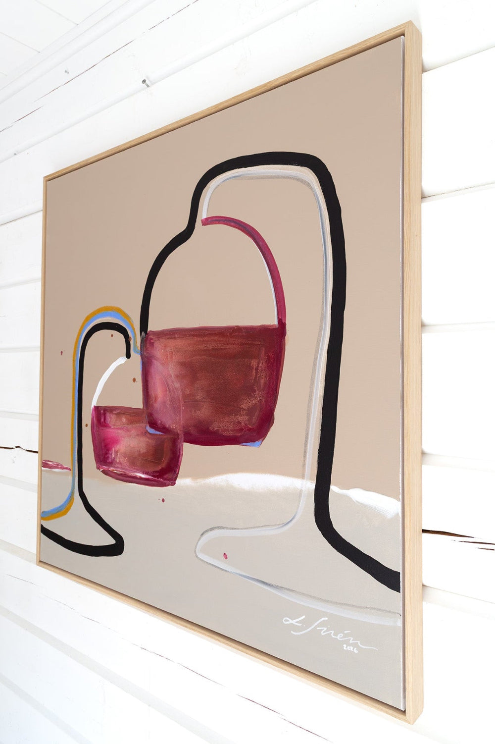 A framed abstract painting featuring two wine glasses with burgundy and pink tones, outlined in black and white, hanging on a white wood-paneled wall by Finnish artist Lotta Sirén