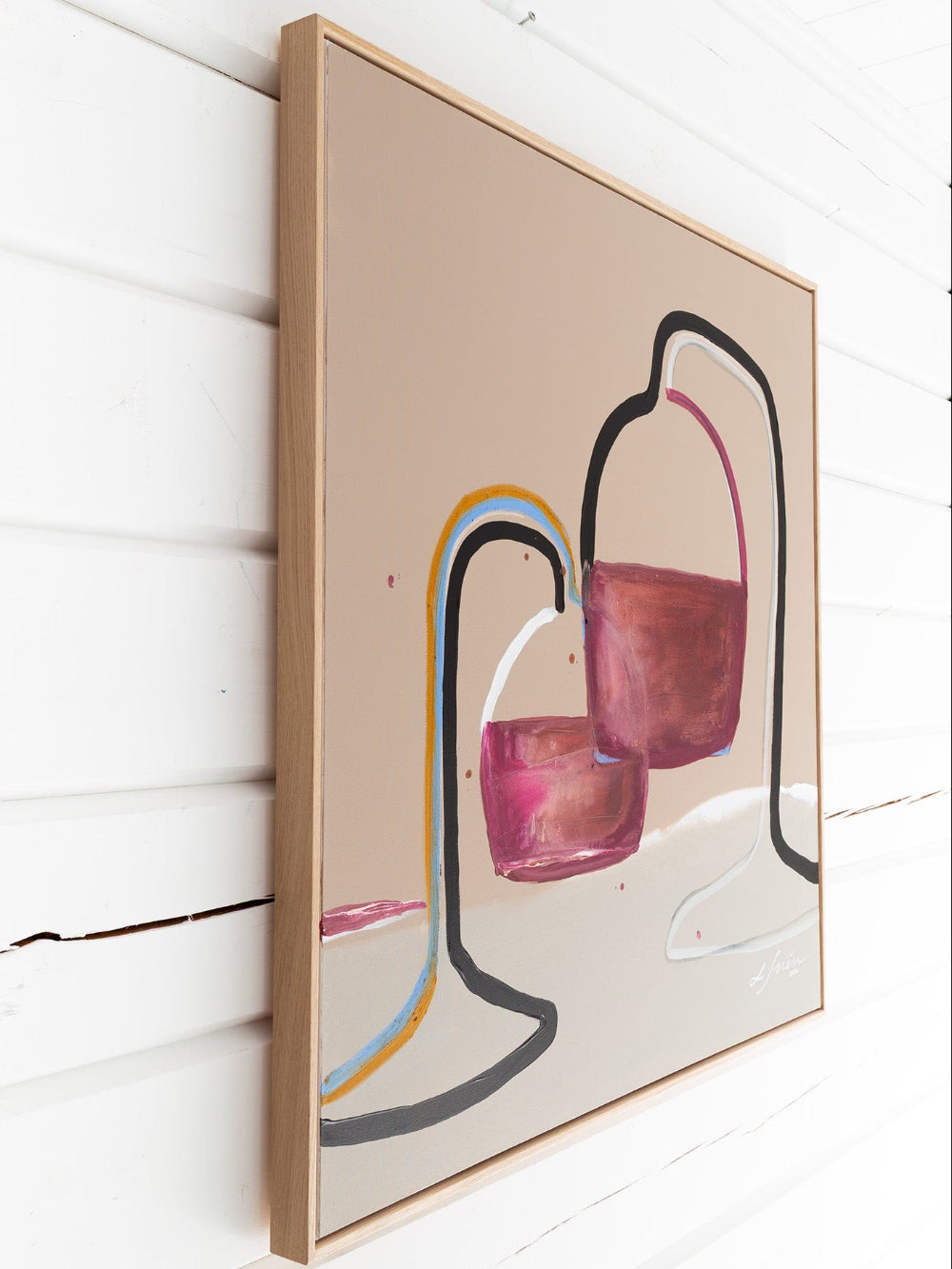 Framed abstract canvas wall art hanging on a white wood-paneled wall. The artwork features abstract shapes in pink, burgundy, black, gray, blue, and yellow on a warm beige background, enclosed in a light natural wood frame by Finnish artist Lotta Sirén