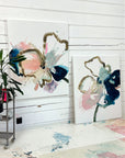 Two abstract floral paintings on a white wall with a plant and cart in the foreground.