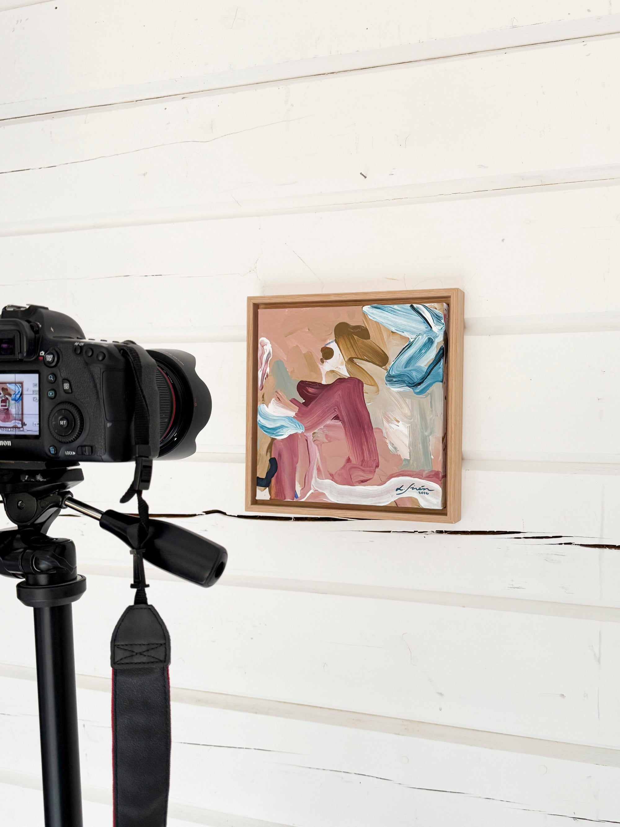 Camera on a tripod capturing a framed abstract painting against a white wooden wall.