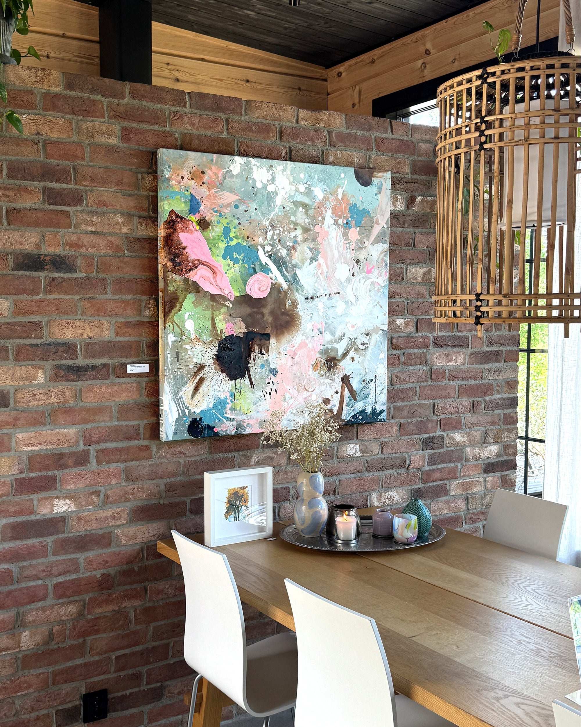 An abstract canvas painting with pink, blue, and white brushstrokes hanging on a rustic red brick wall above a wooden dining table with white chairs in a modern loft setting.