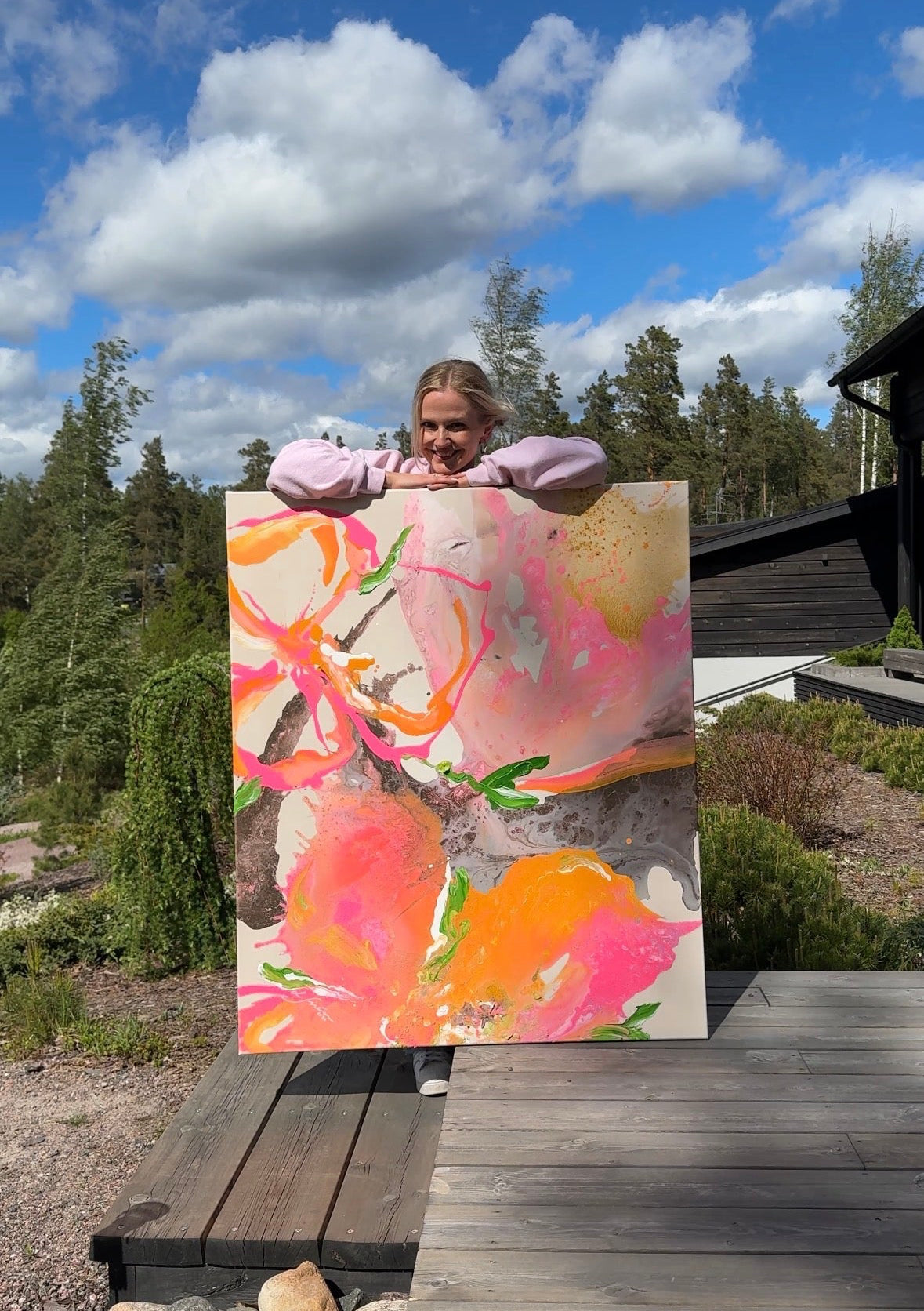 Lotta Sirén holding a colorful semi-abstract floral painting outdoors with trees and sky in the background