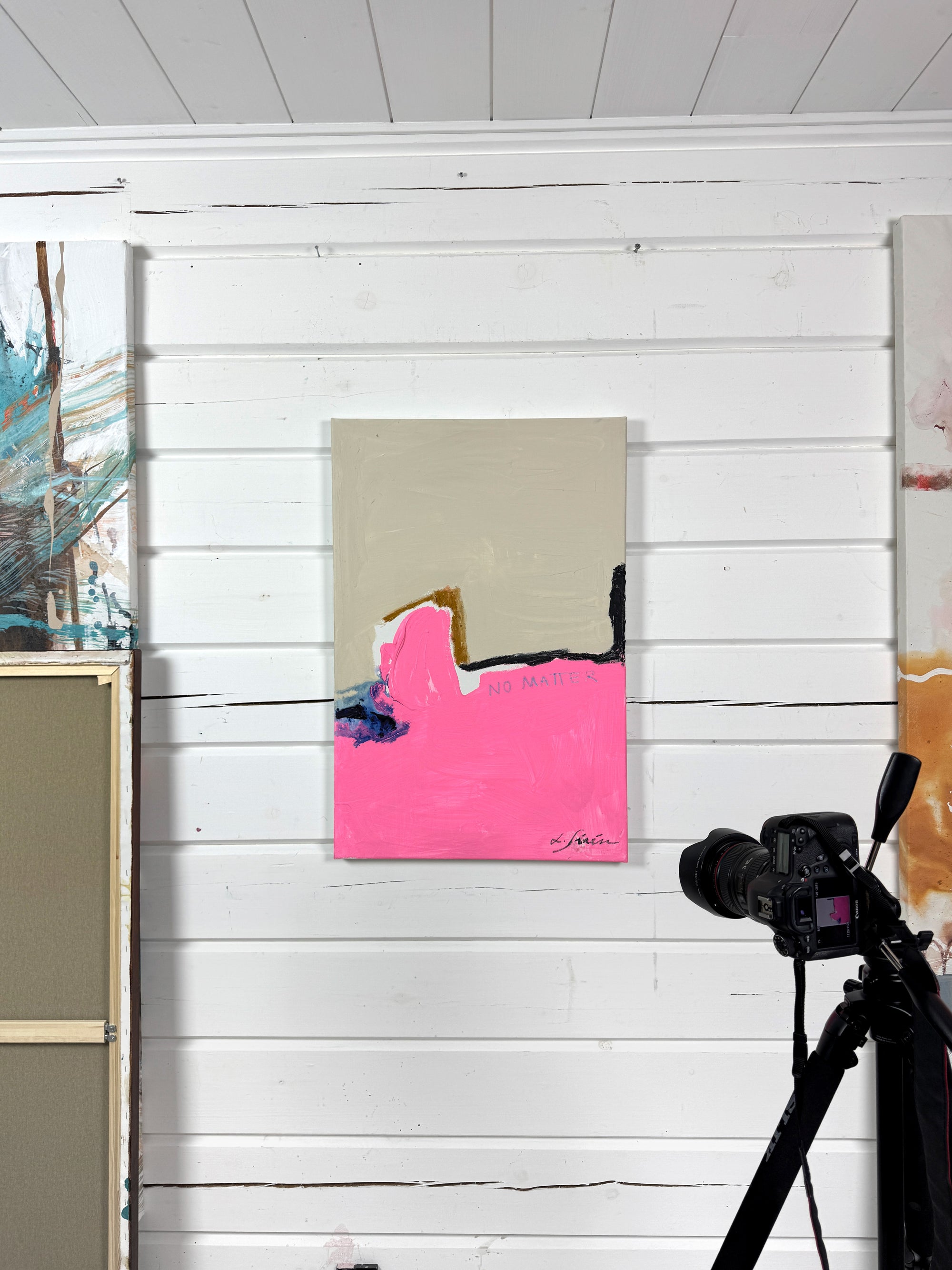 Camera on a tripod and an abstract painting with pink and beige colors against a white wooden studio wall.