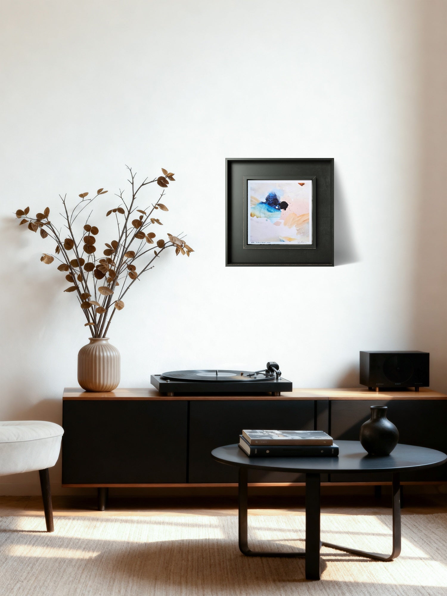 A framed abstract art print with blue, white, and peach tones hangs on a white wall in a modern living room setting above a black media console with a turntable, speaker, and dried flowers.
