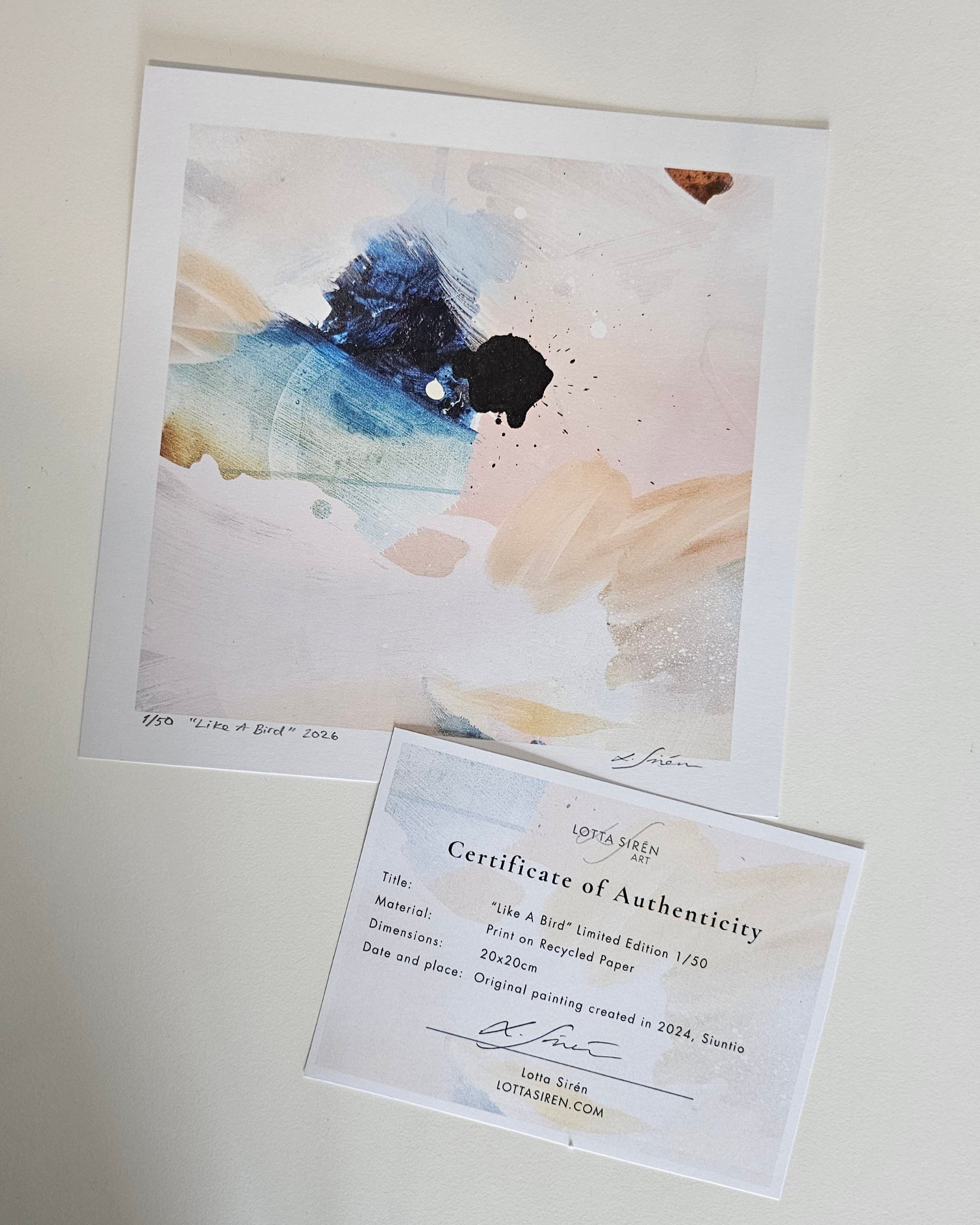 An abstract art print with blue, black, beige, and white brushstrokes, displayed above a Certificate of Authenticity card on a light neutral background.