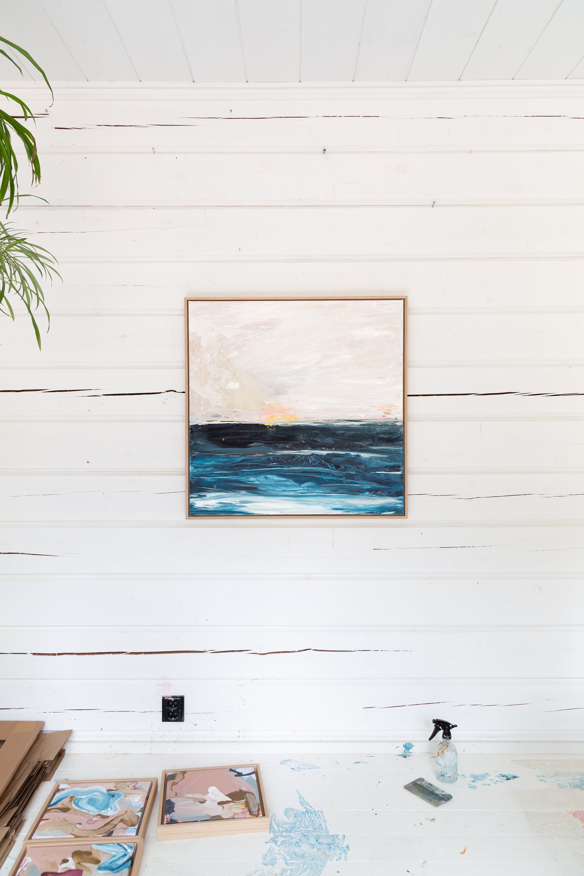 Framed artwork of a seascape on a white wooden wall with art supplies on a table below by Finnish abstract artist Lotta Sirén