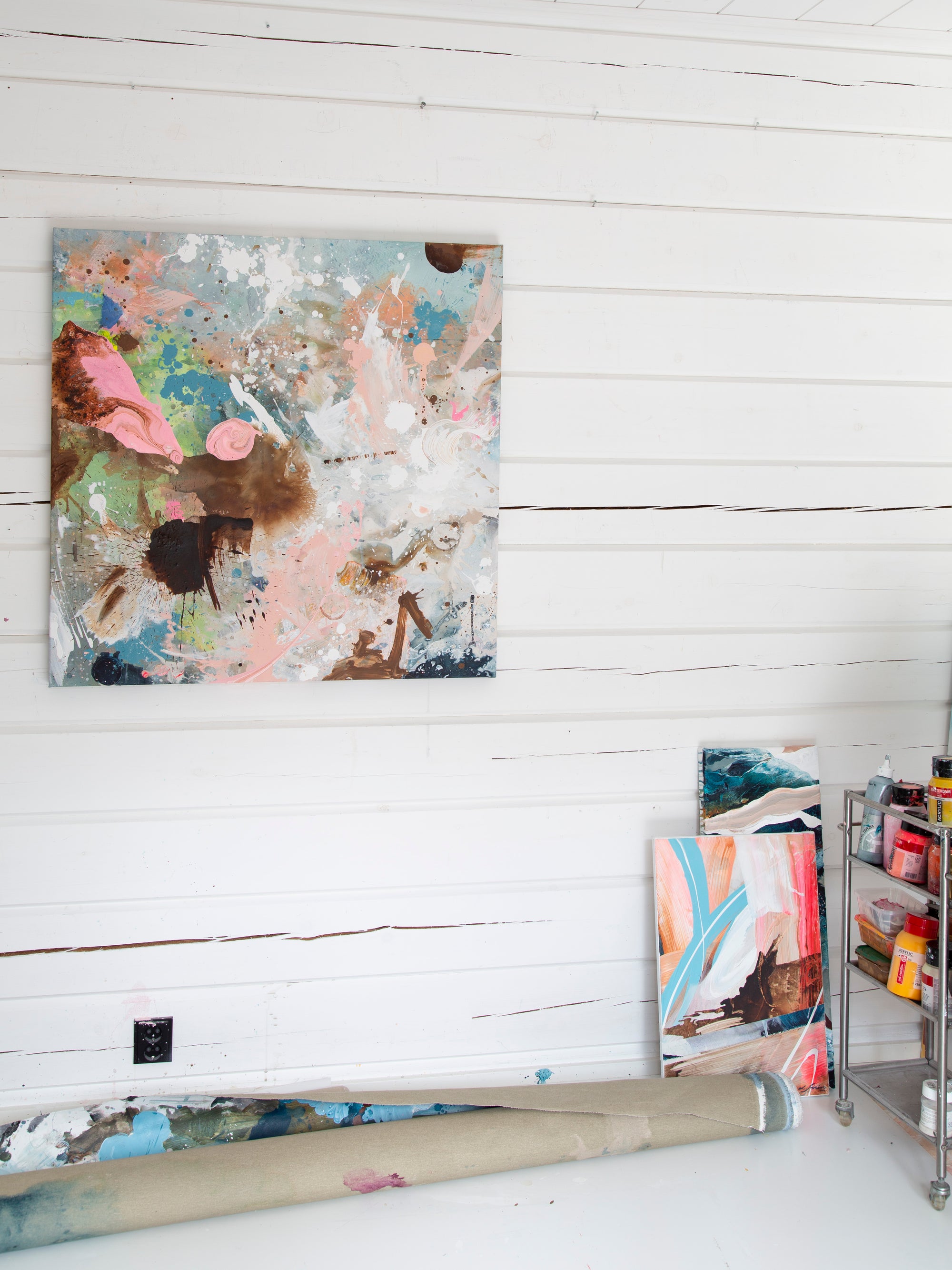 An abstract canvas painting with a multicolor paint-splattered design hangs on a white wood-paneled wall in an artist's studio. A rolled canvas and other art pieces are visible on the floor, along with a metal paint storage cart.